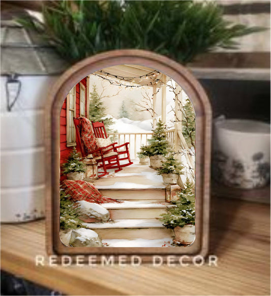 Arched wood-framed canvas art of snowy Christmas porch with red rocking chair and pine trees