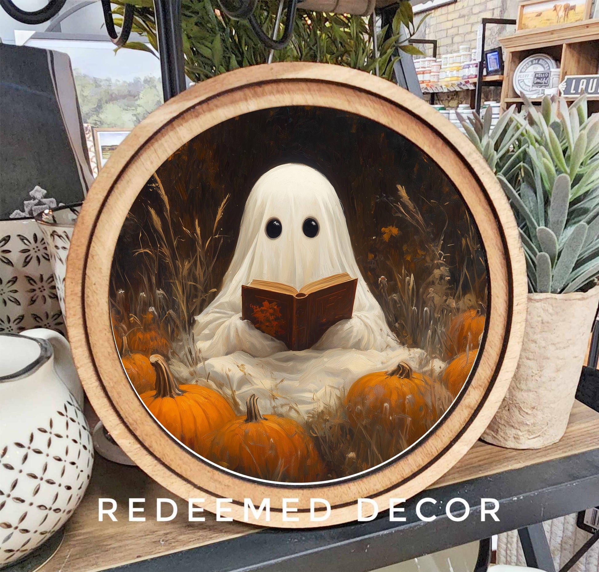 Round wood-framed canvas art of a ghost reading a book among orange pumpkins