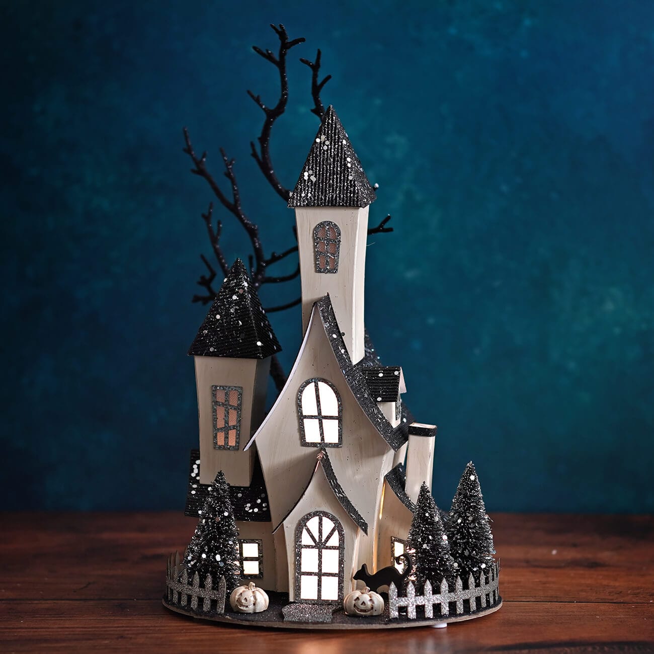 Illuminated cardboard haunted house with black glitter roofs, bare tree, and mini pumpkins on oval base