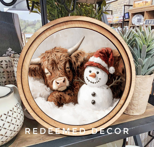 Round wood-framed canvas art of Highland cow with snowman in snow, Christmas theme