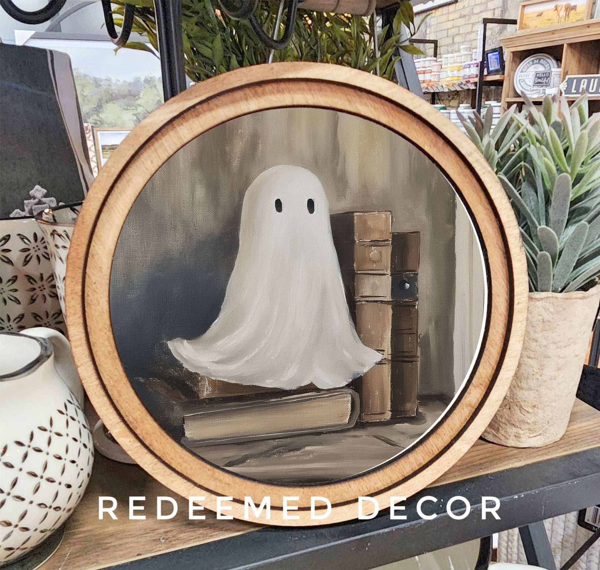 Round wood-framed canvas art featuring a white ghost with old books, neutral tones