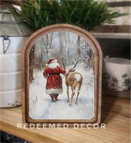 Arched wood-framed canvas art of Santa and reindeer walking in snowy winter forest