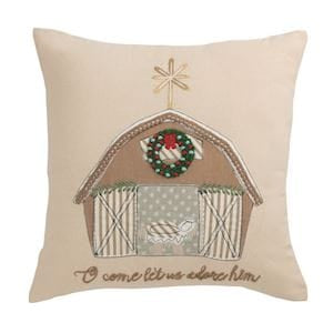 A square beige throw pillow featuring a rustic embroidered barn design with a green wreath, white sheep, and the embroidered phrase 'O come let us adore him' at the bottom.