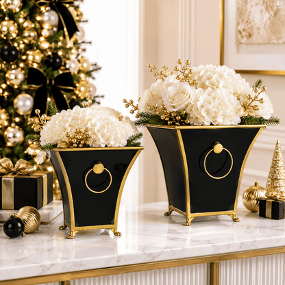 Two black and gold footed cache pots with ring handles filled with white holiday floral arrangements