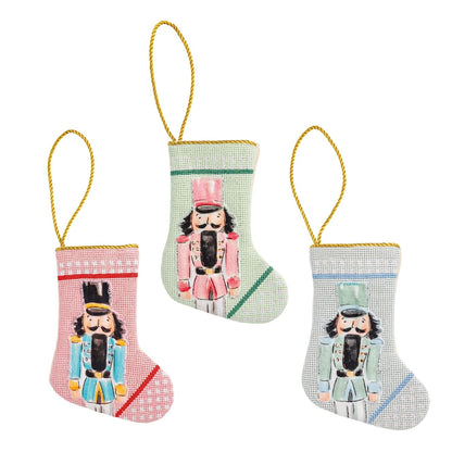Three mini needlepoint nutcracker Christmas stockings in pink, mint, and gray with gold cord loops