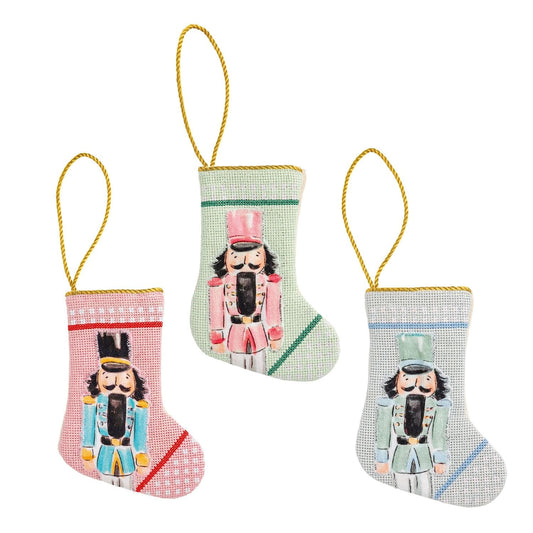 Three mini needlepoint nutcracker Christmas stockings in pink, mint, and gray with gold cord loops