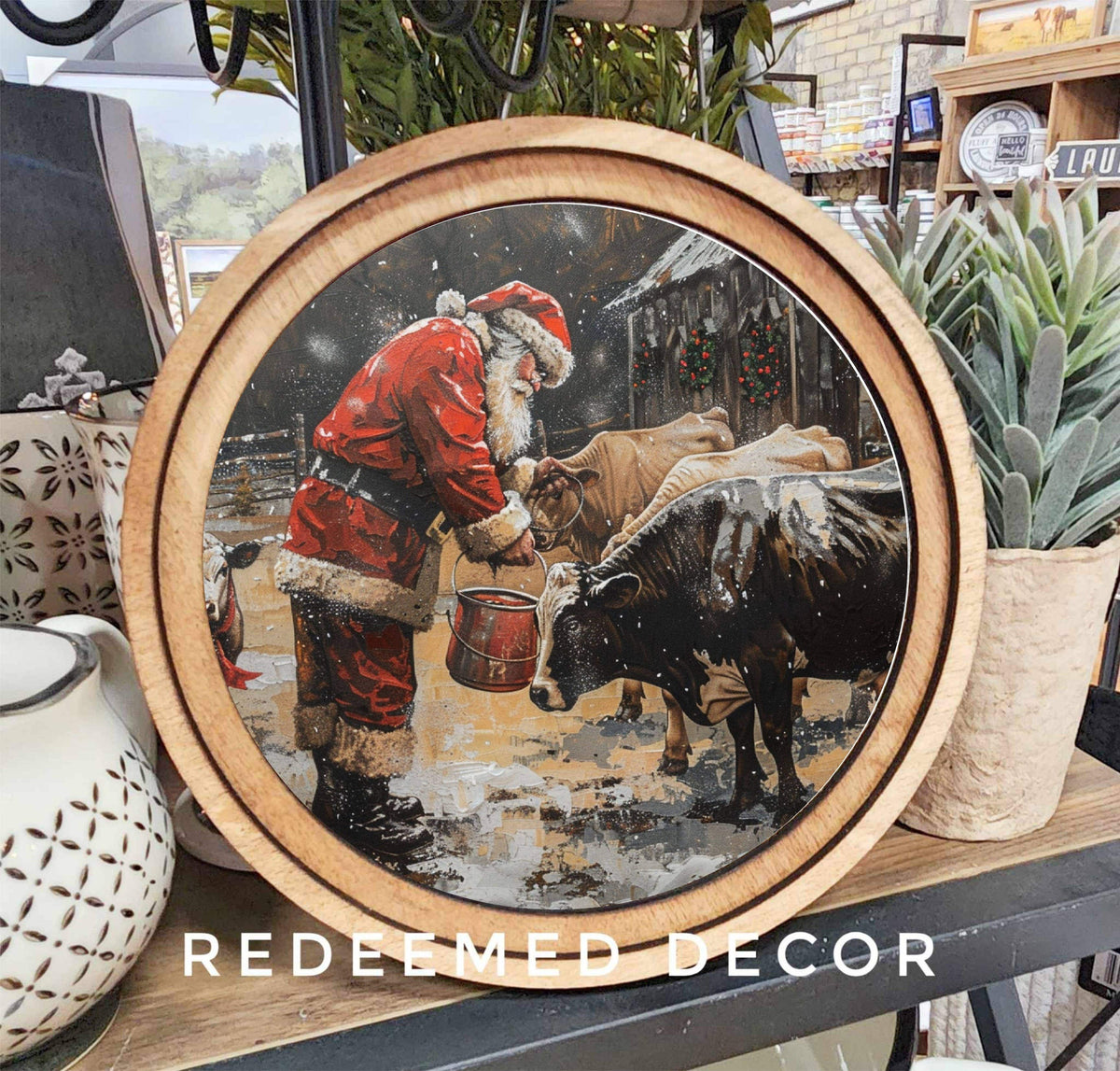 Round wood-framed canvas art showing Santa feeding cows in a snowy barn
