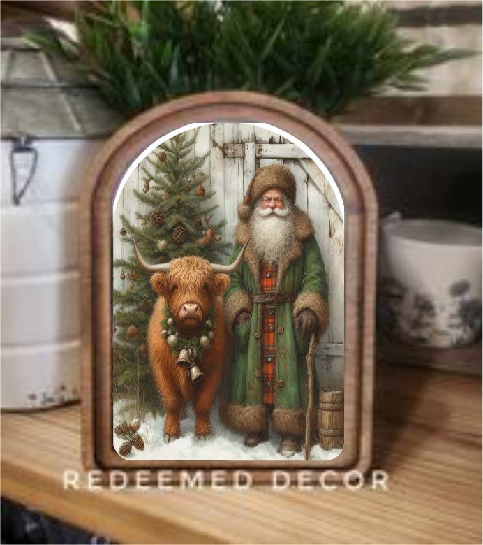 Arched wood-framed art print of Santa in green coat with Highland cow and Christmas tree