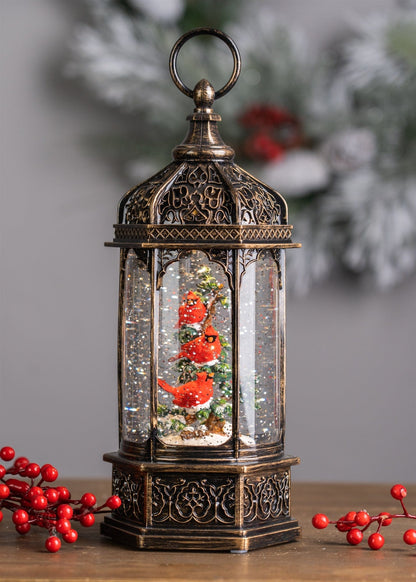 Ornate bronze lantern-style snow globe with two red cardinal birds on a snowy branch inside