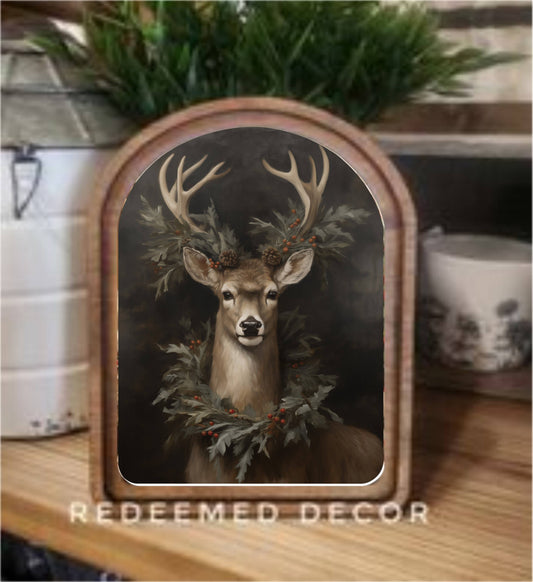 Arched wood-framed canvas art of a deer with antlers and holiday greenery wreath