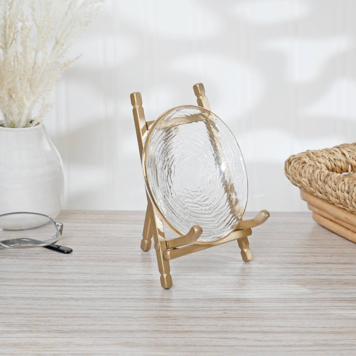Gold satin metal display easel holding a clear glass decorative plate on a wooden surface