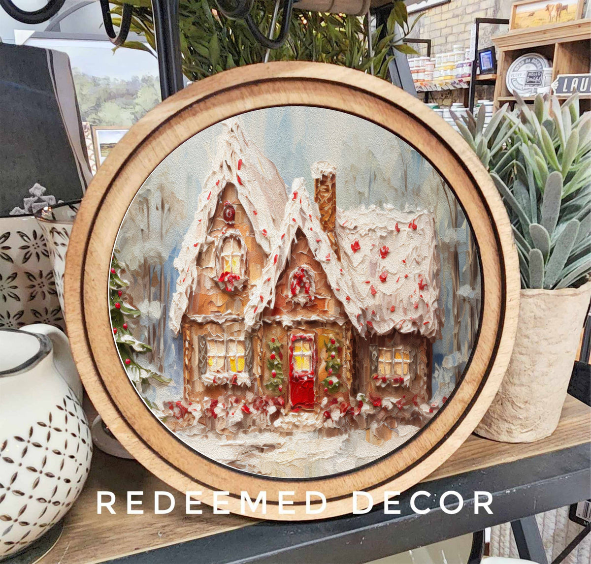 Round wood-framed canvas art featuring a snow-covered gingerbread cottage with red door