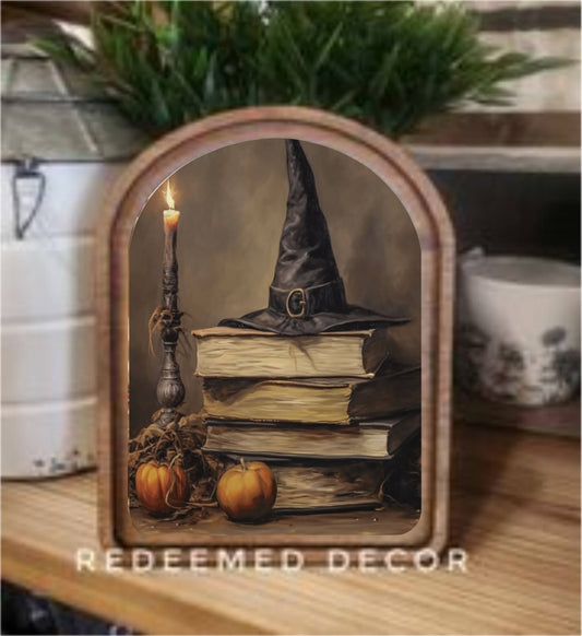 Arched wood-framed Halloween canvas art with witch hat, stacked books, candle, and pumpkins