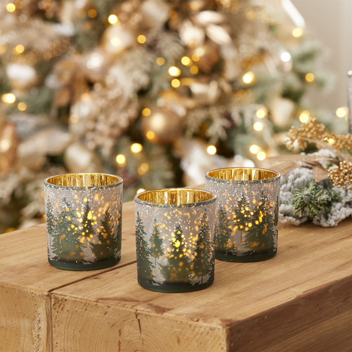 Set of three frosted glass votive holders with winter forest design, glowing gold interiors, on wood surface