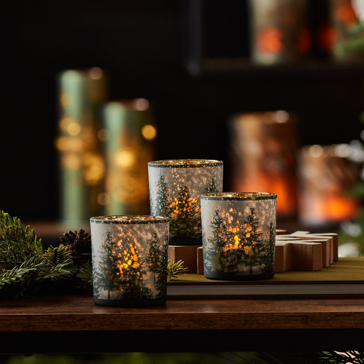 Set of three frosted glass votive holders with winter forest scene, glowing with candlelight