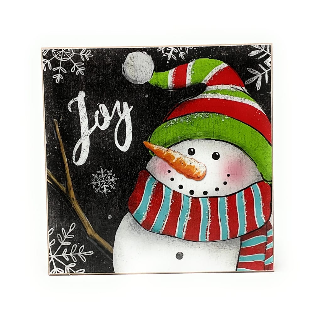Square wood block sign with cheerful snowman wearing striped hat and scarf, 'Joy' text on black background