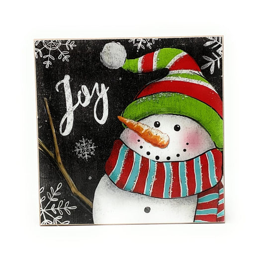 Square wood block sign with cheerful snowman wearing striped hat and scarf, 'Joy' text on black background