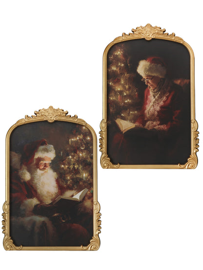 Pair of ornate gold-framed paintings depicting Santa and Mrs. Claus reading books by a Christmas tree