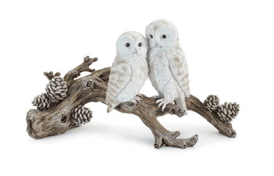 Two white owl figurines perched on a weathered branch with pinecones