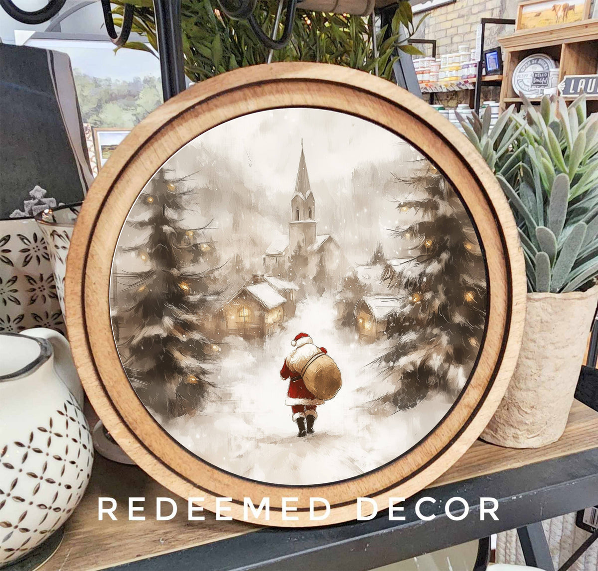 Round wood-framed canvas art of Santa walking toward snowy village with pine trees