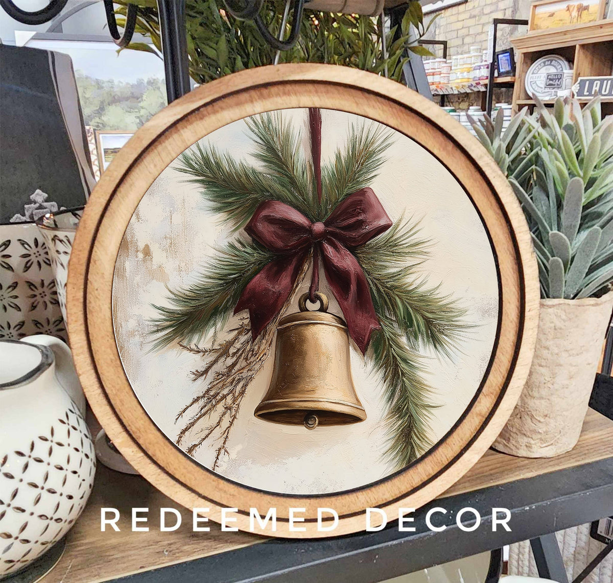 Round wood-framed canvas art with gold bell, pine sprigs, and burgundy bow