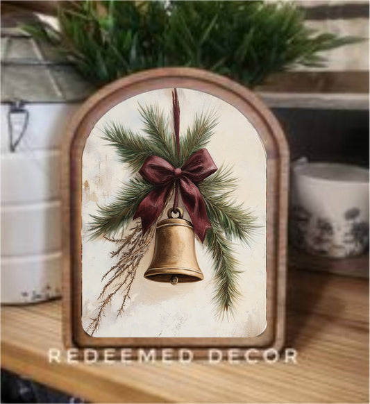 Arched wood-framed canvas art depicting a gold bell with red bow and pine branches