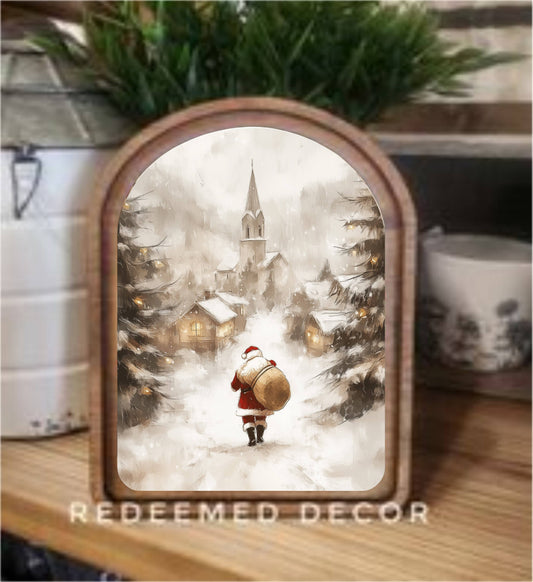 Arched wood-framed canvas art of Santa walking snowy village toward church steeple
