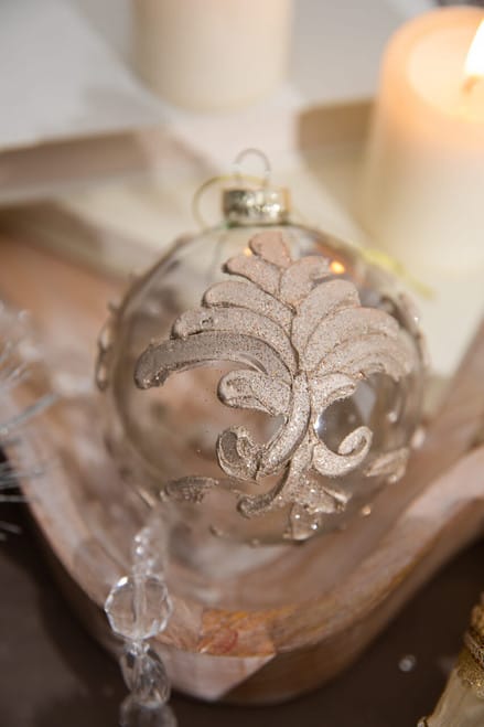 Clear glass ball ornament with gold glitter acanthus leaf design, displayed on wooden tray with candles